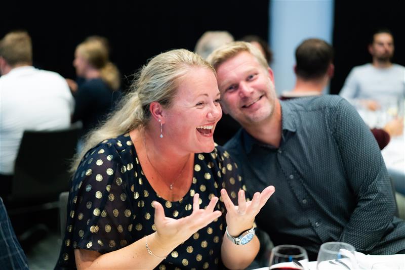 Foto av to folk på konferansen, med flere i bakgrunnen av bildet med uskarpt fokus. De to personene i forgrunnen ler og har det gøy.